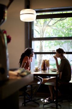 two women sitting at a table in a restaurant eating pizza and drinking coffee, looking out the window