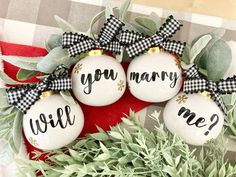 three christmas ornaments with the words you marry, will me? on them next to some greenery