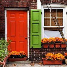 an orange door with green shutters and potted flowers on the side of a brick building