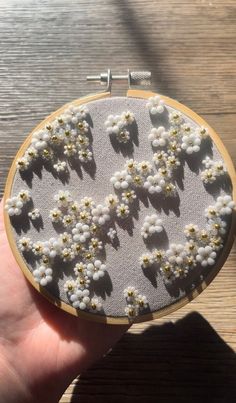 a hand holding a small embroidery project with white and yellow flowers on grey fabric in front of a wooden table