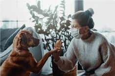 a woman wearing a face mask sitting on a couch petting a dog's paw