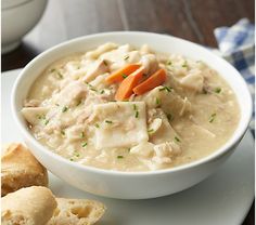 a bowl of chicken and dumpling soup on a white plate next to two rolls