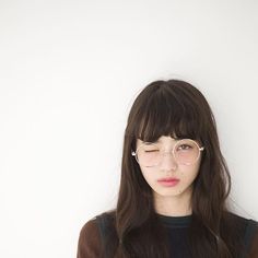 a woman with glasses and long hair standing in front of a white wall looking at the camera