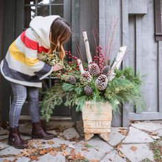 a woman placing pine cones on top of a planter filled with evergreen and berries