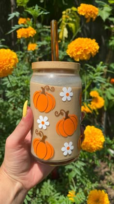 a hand holding a mason jar with orange and white pumpkins on it in front of yellow flowers