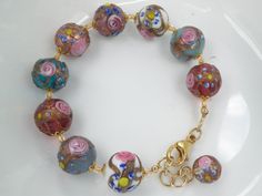 "The Fiorato (flower) beads, that some people call \"wedding cake beads\", on this bracelet, are Vintage Murano Glass Beads. This type of handmade, lamp worked Murano glass, Venetian Beads, are some of the first beads I ever bought in Venice, Italy. The assortment of 14-15 mm, round beads were made one-by-one under the flame. The glass maker starts with a Murano glass cane (or rod) to make the base color, and then adds an Aventurina (copper filings), band around the center of the bead. Next the Multicolor Beaded Murano Glass Jewelry, Handmade Vintage Jewelry In Murano Glass, Elegant Round Murano Glass Necklace, Pastel White, Vintage Murano Glass Beaded Necklaces, Handmade Lamp, Bohemian Murano Glass Beads, Colorful, Murano Glass Beads, Flower Beads