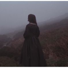 a woman standing on top of a lush green hillside in front of a foggy sky