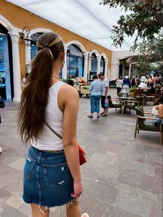 a woman with long hair standing on the sidewalk in front of a building and people walking around