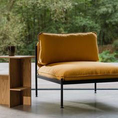 a chair with a yellow cushion next to a small side table and trees in the background