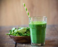 a green smoothie in a glass next to a bowl of spinach and a straw