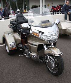 a silver motorcycle parked in front of a white building with people standing around the area