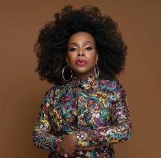 a woman with an afro standing in front of a brown background wearing a colorful dress