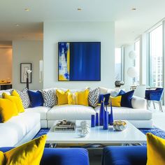 a living room filled with white couches covered in blue and yellow pillows next to tall windows