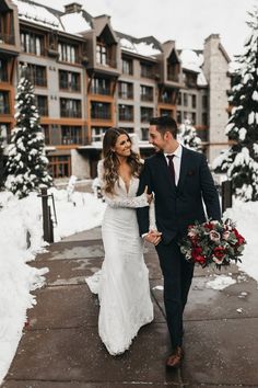 a bride and groom walking through the snow in front of an apartment building holding hands