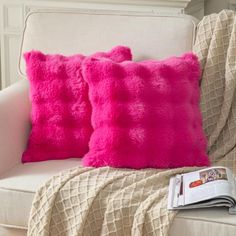 two pink pillows sitting on top of a white chair next to a book and blanket