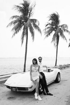 a man and woman sitting on the hood of a sports car in front of palm trees