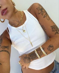 a woman with tattoos on her arms and chest wearing a white tank top, blue jeans and gold chain bracelets