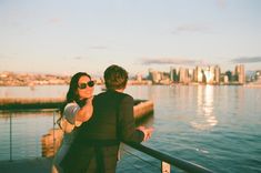 a man and woman are standing on a pier looking at the water with city in the background