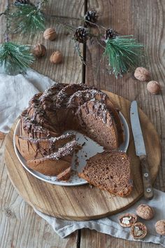 a chocolate bundt cake on a wooden plate with one slice cut out and some nuts around it