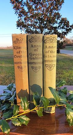 three books sitting on top of a wooden table next to a leafy plant in front of a tree