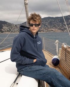 a man sitting on the deck of a sailboat wearing sunglasses and a hoodie