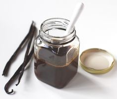 a jar filled with liquid next to two spoons on a white tablecloth and some vanilla sticks