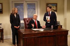 two men and a woman standing in front of a wooden desk with papers on it