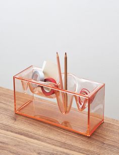 an orange plastic container with pens and pencils in it on a wooden table next to a white wall