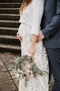 a pregnant woman in a white dress holds her husband's hand as they stand next to some steps