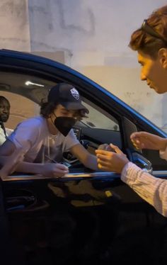 two men sitting in a car and one is handing something to another man who is wearing a face mask