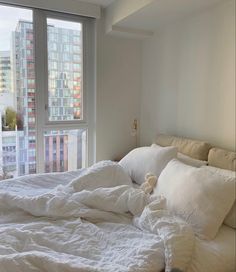 an unmade bed with white sheets and pillows in front of a large window overlooking the city