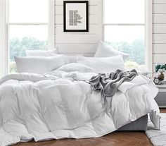 a bed with white comforters and pillows in a room next to two large windows