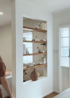 a woman standing in front of a white wall with shelves filled with books and vases