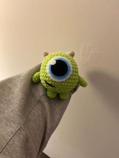 a green stuffed animal with big eyes sitting on someone's arm in front of a white wall