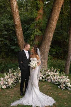 a bride and groom standing in front of some trees with flowers on the ground next to them