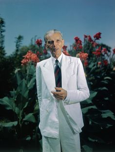 an older man in a white suit and tie standing next to some red flower bushes