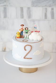 a birthday cake with toy figures on top and clouds in the background, sitting on a white table