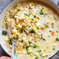 a person is holding a spoon in a bowl of soup with corn and zucchini