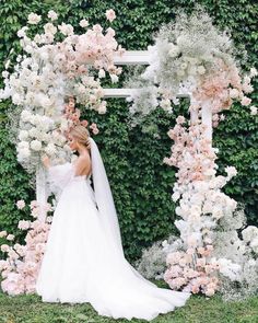 a woman in a wedding dress standing under a flower covered arch with white and pink flowers