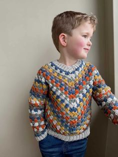 a young boy standing next to a window wearing a colorful knitted sweater and jeans