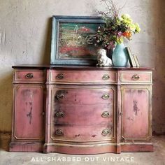 an old dresser with flowers and a painting on it's sideboard in front of a wall