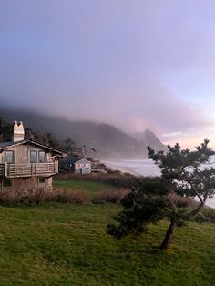 a house sitting on top of a lush green hillside next to the ocean with fog in the air