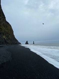 a bird flying over the ocean next to a rocky cliff on a beach with waves coming in