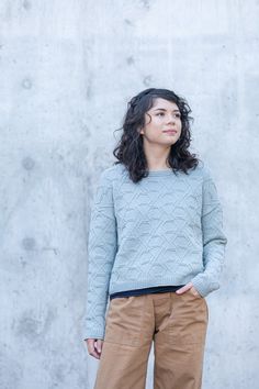 a woman standing in front of a concrete wall wearing brown pants and a blue sweater