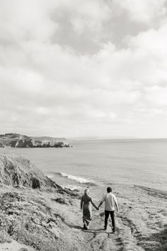 two people holding hands while walking down a path near the ocean on a cloudy day