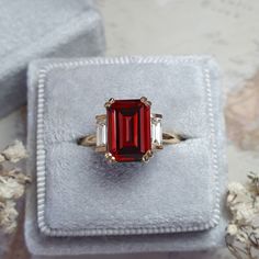 a red and white diamond ring sitting on top of a velvet box with flowers in the background