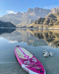 a pink and white surfboard sitting on the shore of a lake with mountains in the background