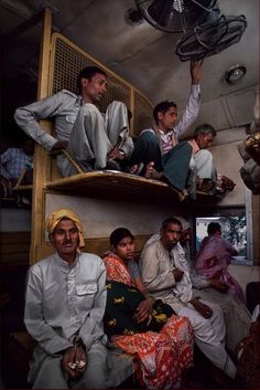 several people sitting on bunk beds in a small room with fans hanging from the ceiling