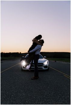 a man and woman kissing in front of a sports car on the road at sunset