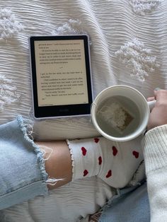 a person sitting on a bed with their legs crossed holding a coffee cup and an electronic device